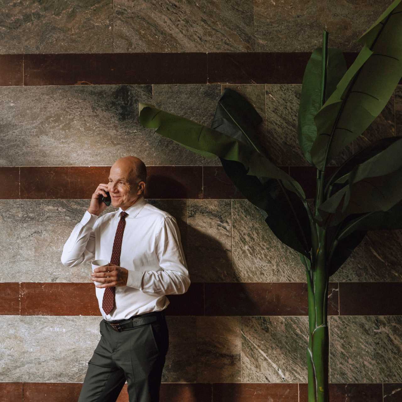 Man in a white button up shirt and red tie on phone standing in front of wood and granite striped wall with large plant to his right-hand side.