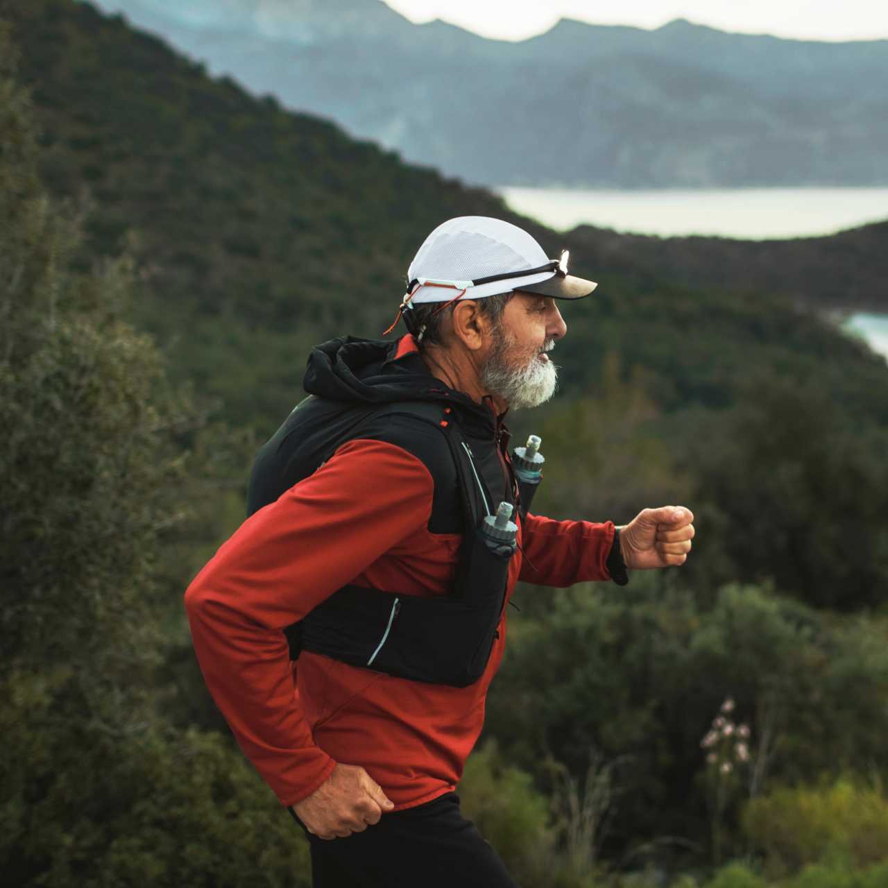 Homme d’un certain âge portant un survêtement rouge, une veste de course noire et une casquette blanche qui fait un trail dans la montagne