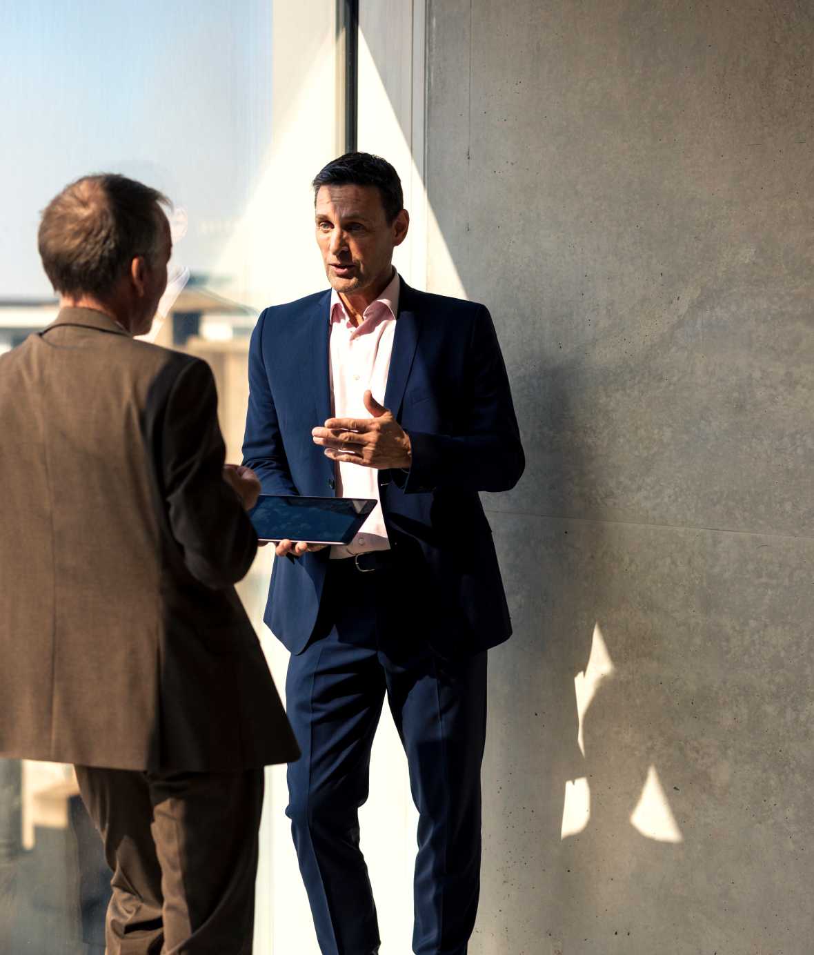 Two colleagues in suites standing in front of full length window, talking and holding smart tablet.