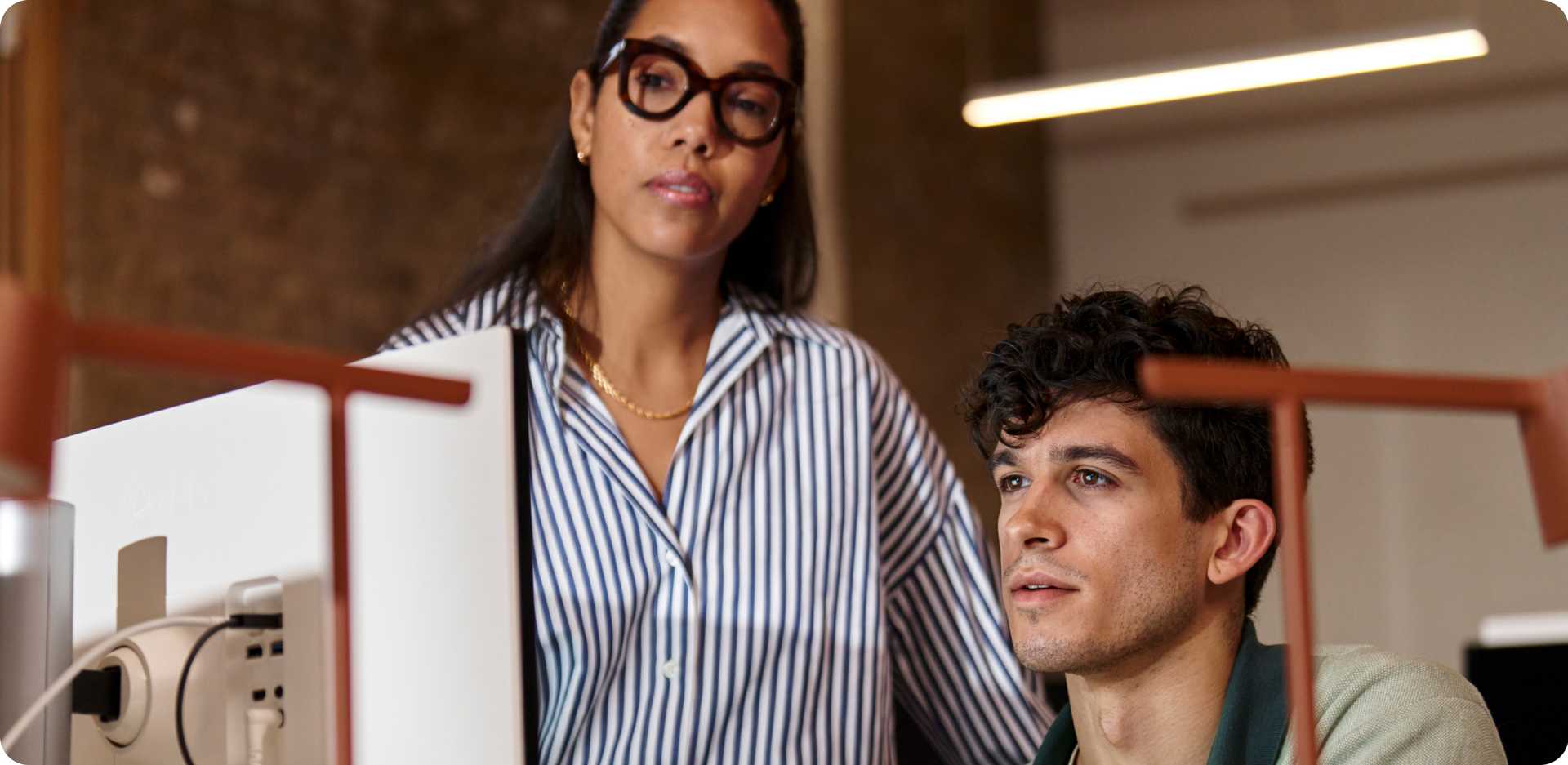 A woman wearing a striped blouse and glasses stands beside a man seated at a desk, looking at a computer.