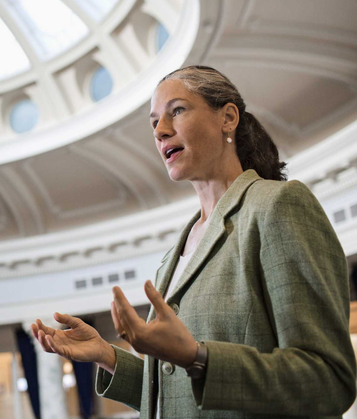 Woman in green blazer speaking and moving her hands inside of a government building