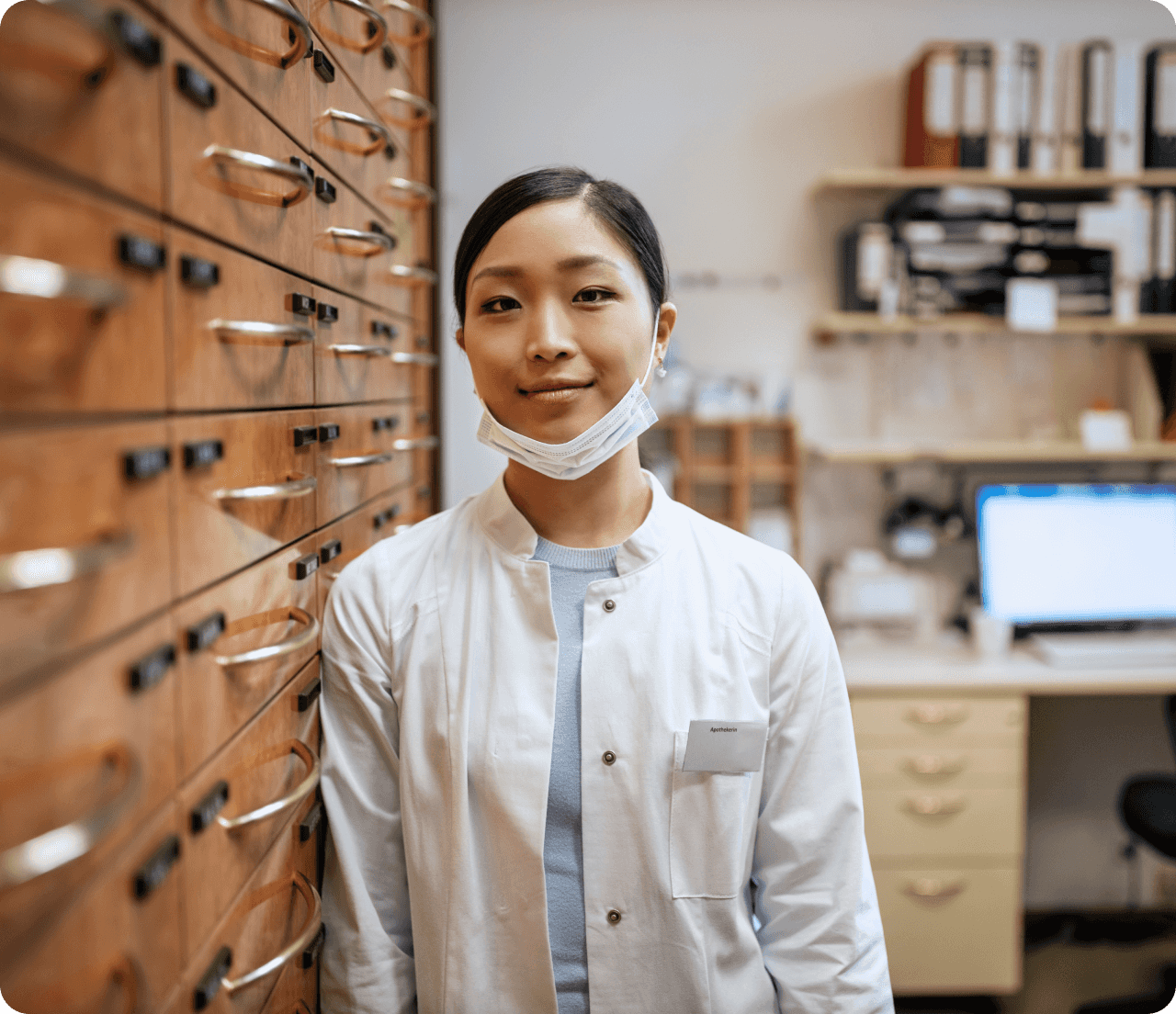 Female healthcare worker in white coat and mask pulled down standing in office and smiling at camera.