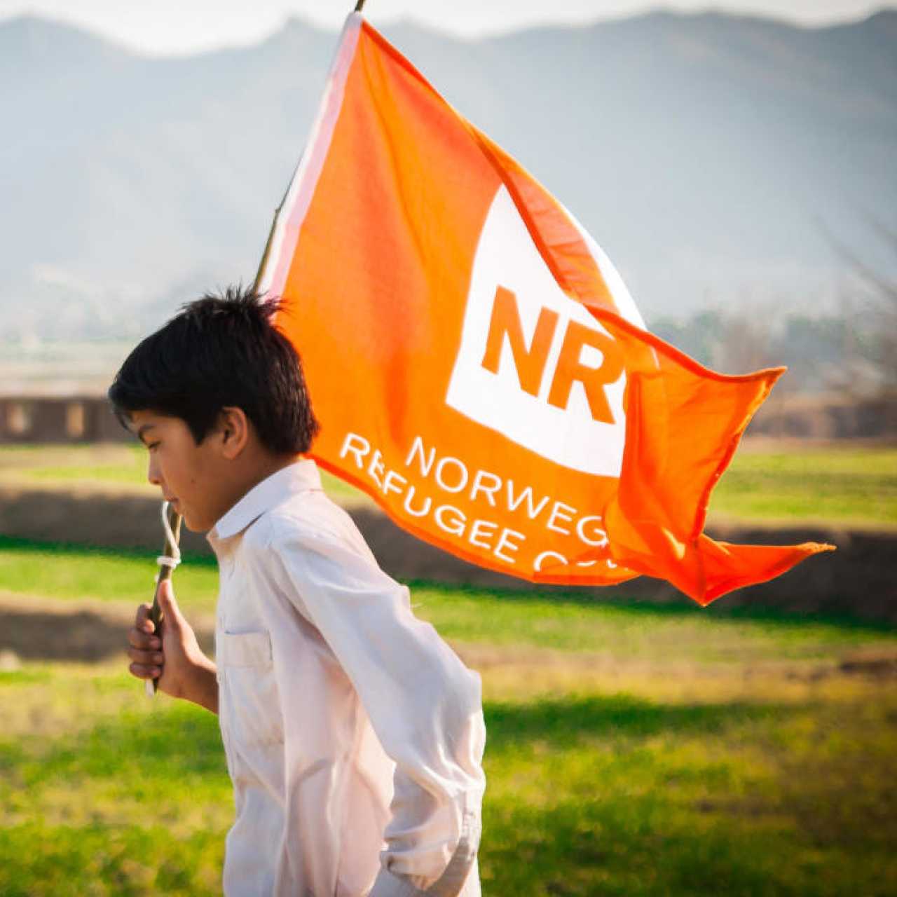 Ein Kind läuft auf einer grünen Wiese und hält eine orangefarbene Flagge der Norwegischen Flüchtlingshilfe in der Hand