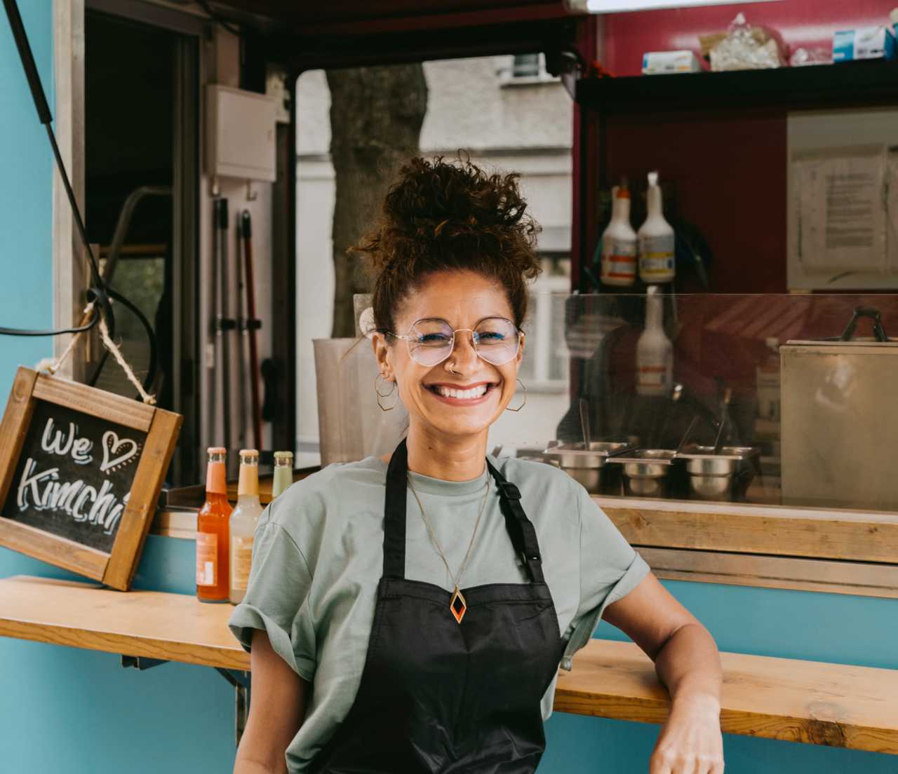 Young woman in glasses and an apron smiling and standing in front of a food stand.