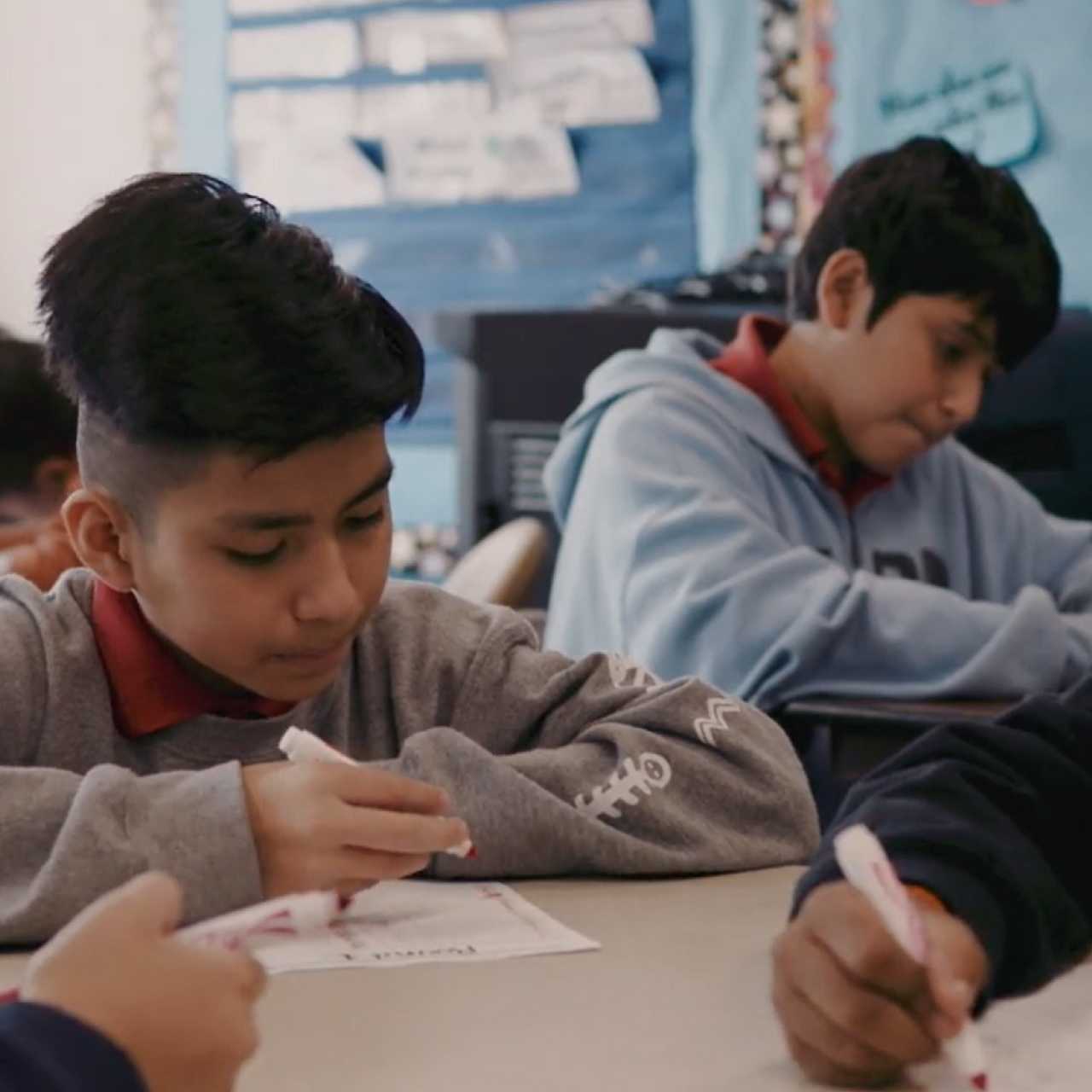 Deux petits garçons assis derrière des bureaux dans une salle de classe d’une école primaire, qui écrivent sur du papier avec des marqueurs