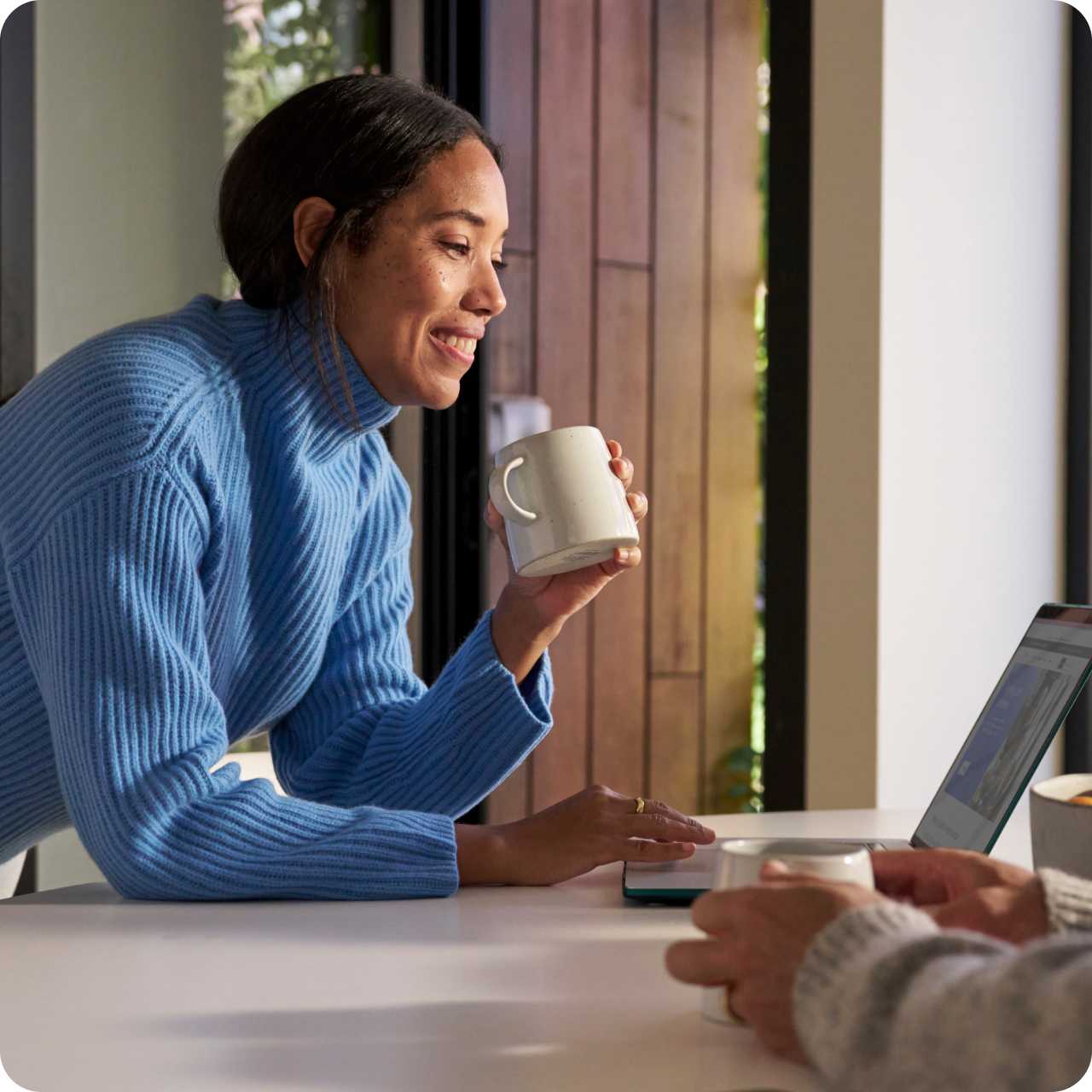 Image of a woman leaning over a table working on her laptop while holding a mug. 