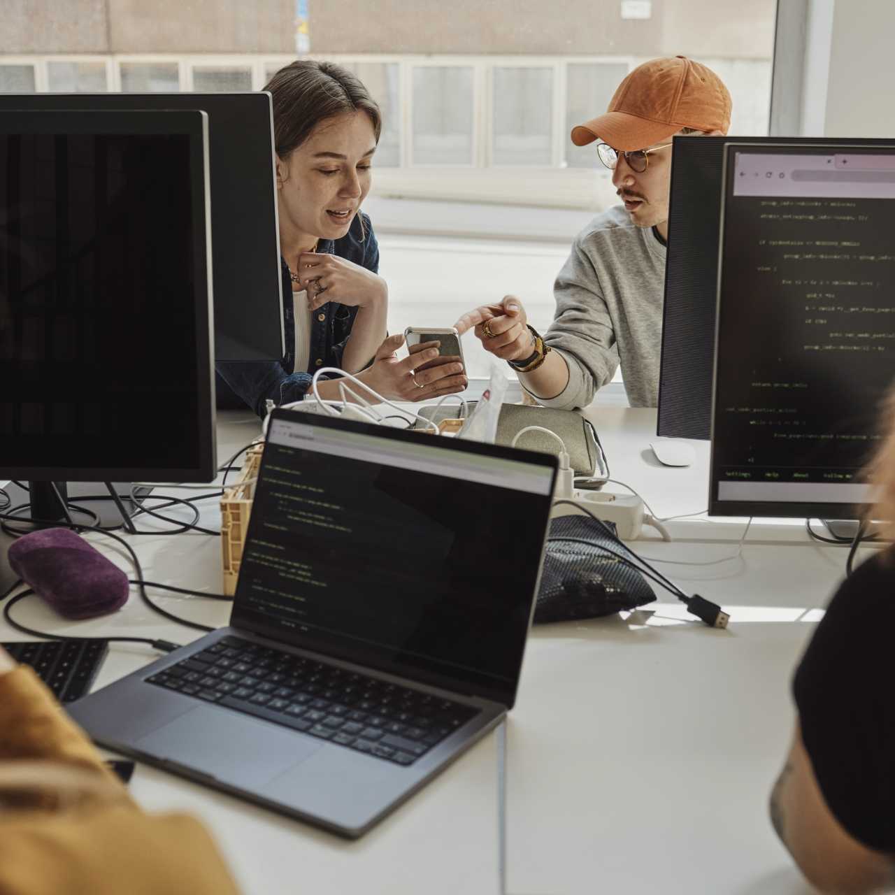 Image de collègues assis devant leurs ordinateurs, regardant le smartphone de la collaboratrice. 