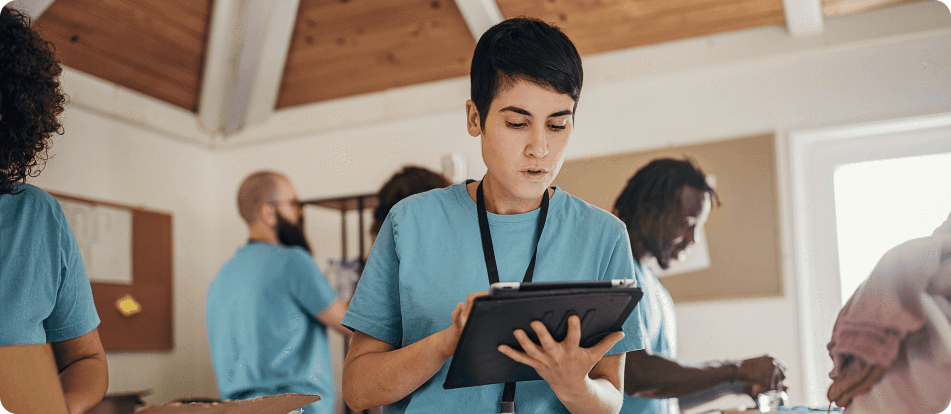 Woman in blue shirt wearing lanyard while looking down at smart device and standing in room with people wearing the same uniform