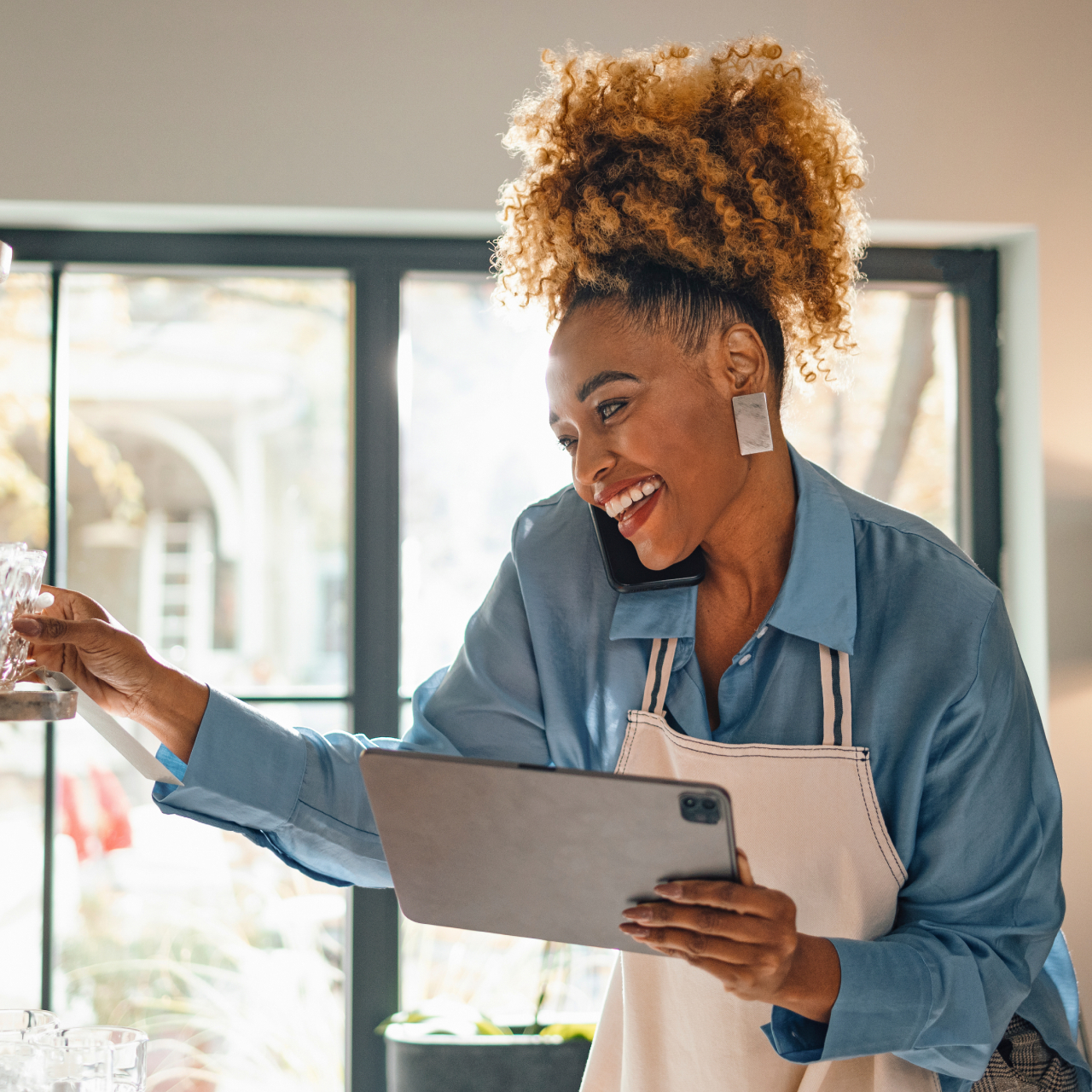 Image of a smiling woman wearing an apron, holding a phone to her ear while also holding a smart device.