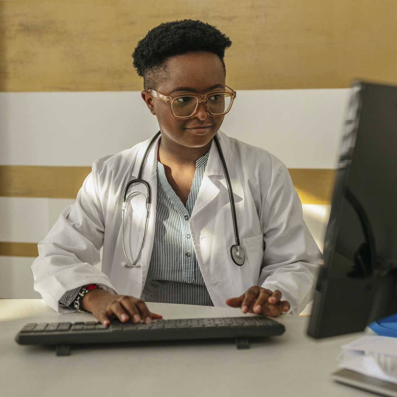 A doctor in white coat and a stethoscope around neck sitting at desk and typing on desktop computer