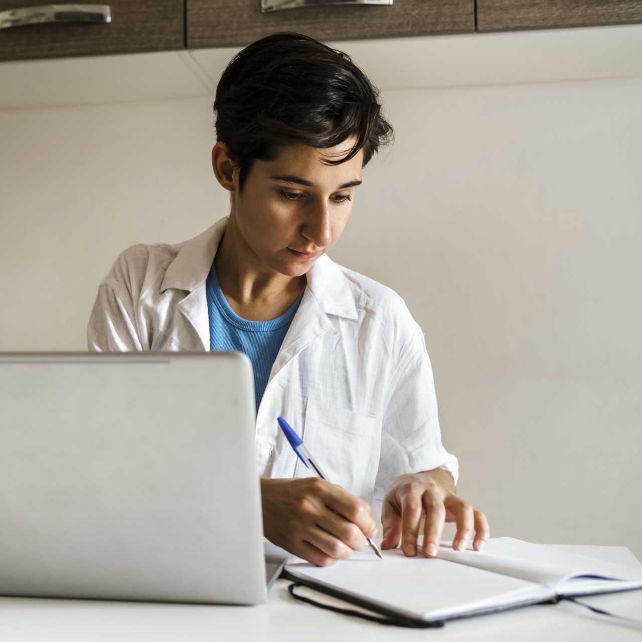 Healthcare worker in white coat sitting at desk with laptop and writing in notebook with pen