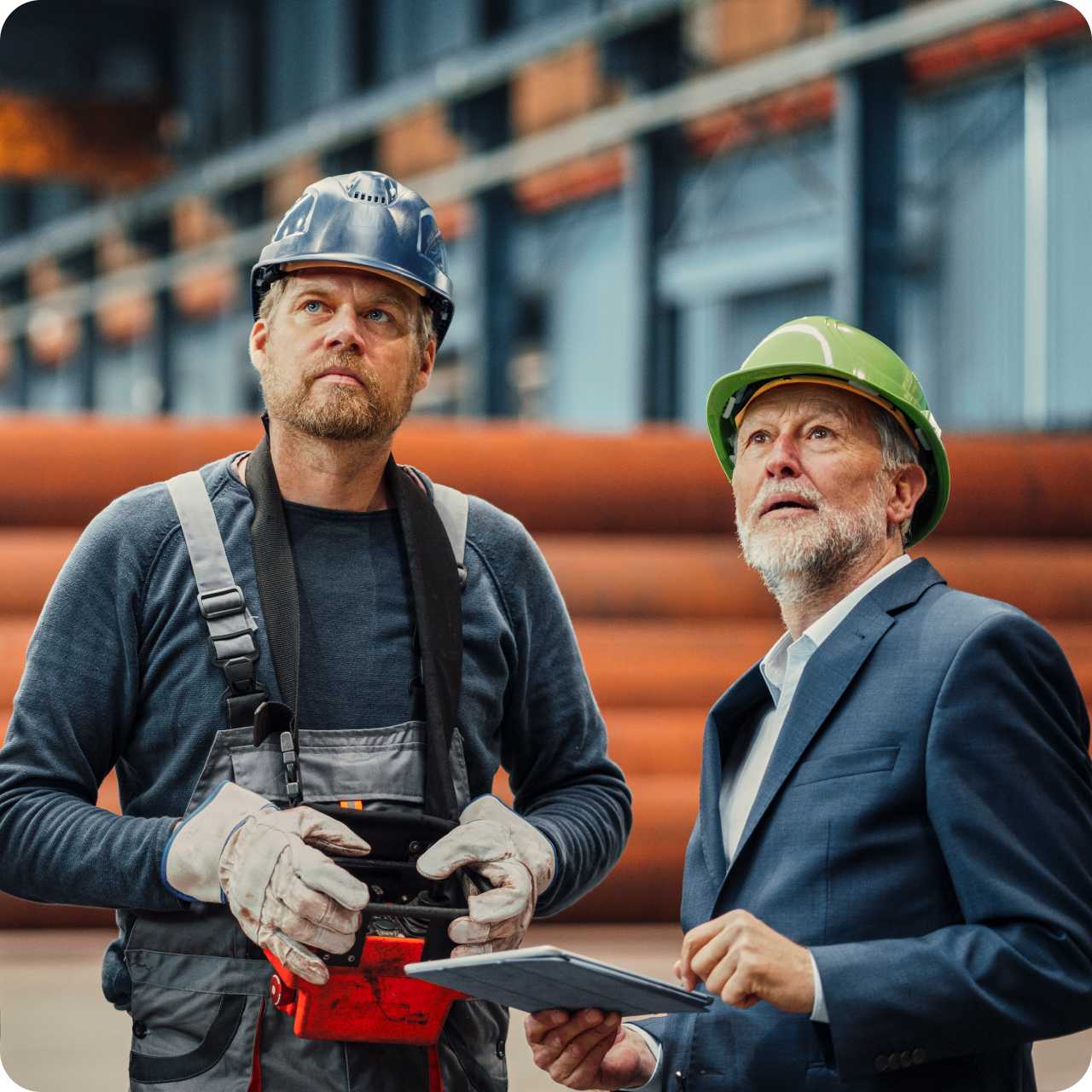 A manager and worker wearing hard helmets view and inspect a production floor. 