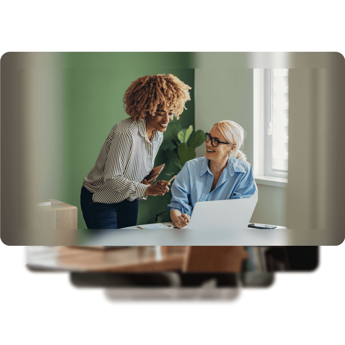 An image of two women working on a laptop at a desk, with a green wall and a plant in the the background.