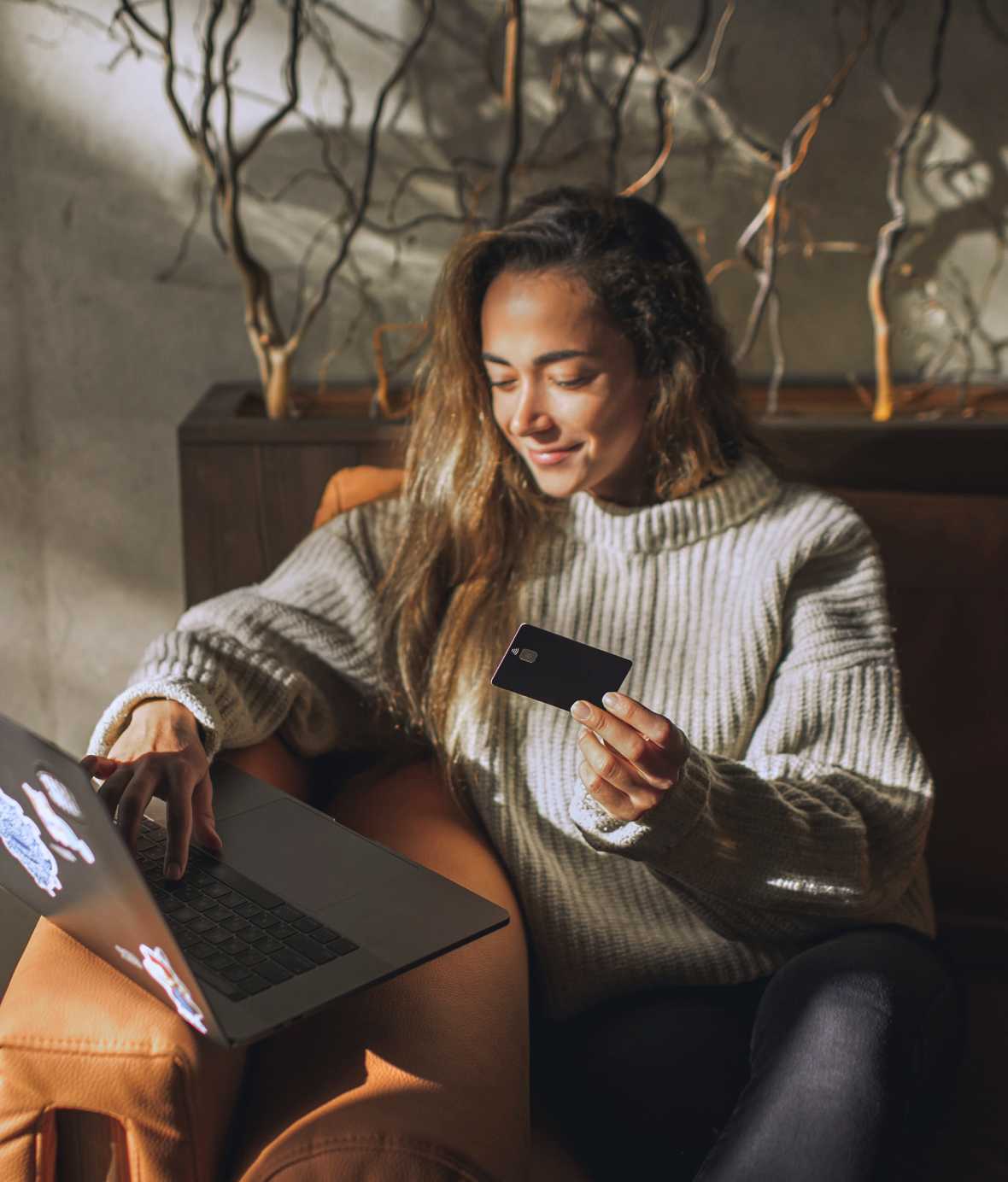 Young woman in sweater sitting on couch with laptop out and credit card in hand.