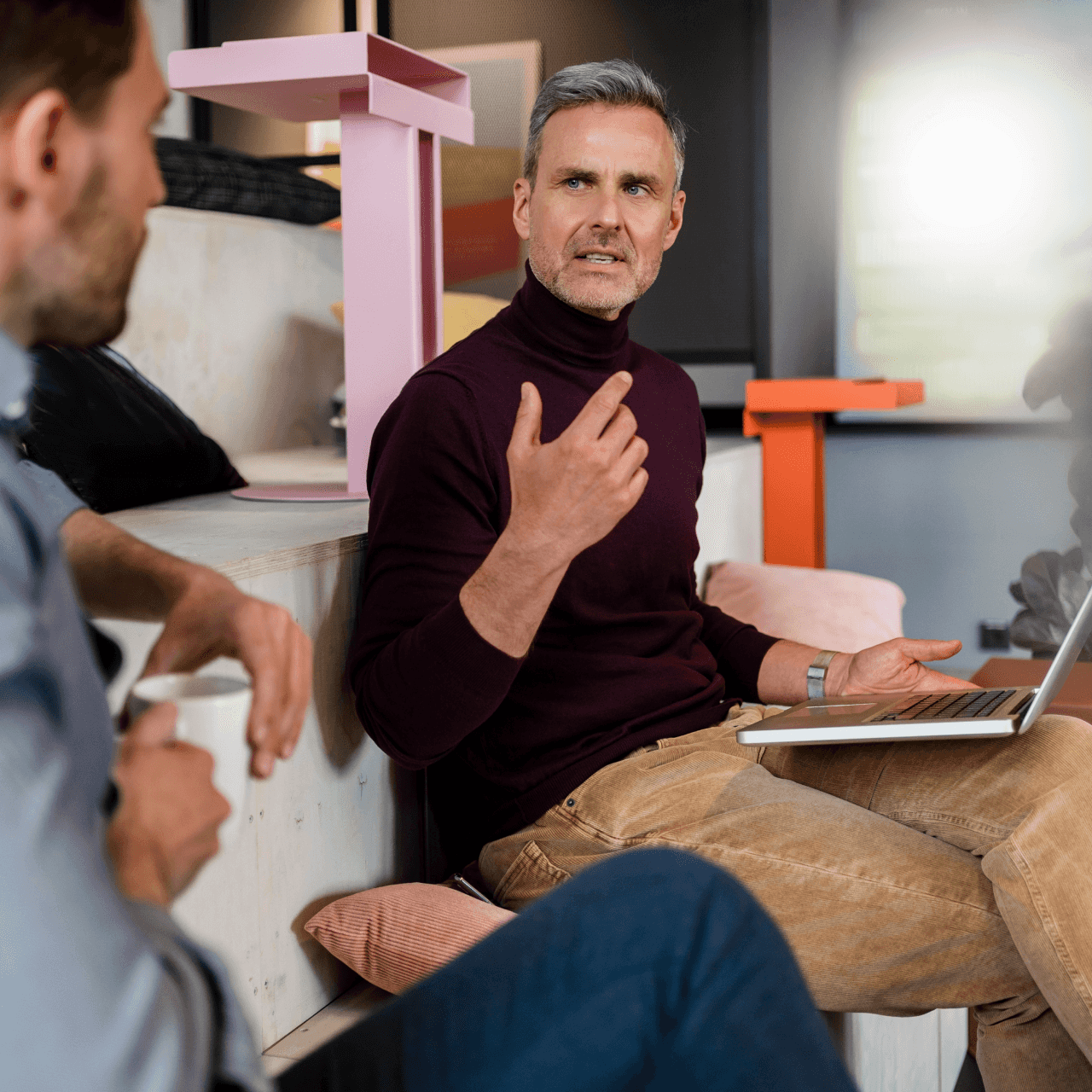 Image of two men sitting in the office. One with a cup of coffee and the other with a laptop open.