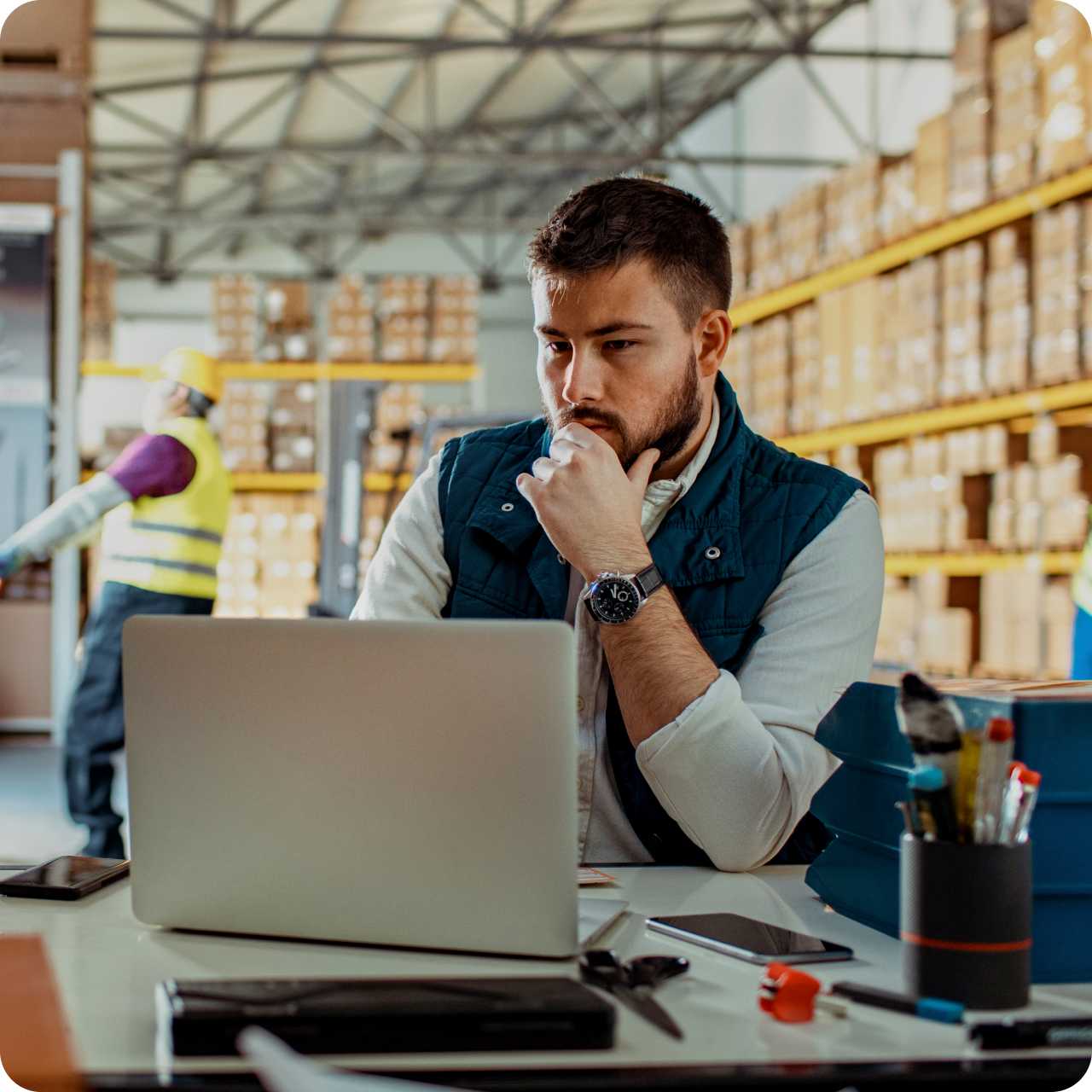 A man sits at a desk and uses a laptop on the factory floor while a warehouse worker works in the background. 