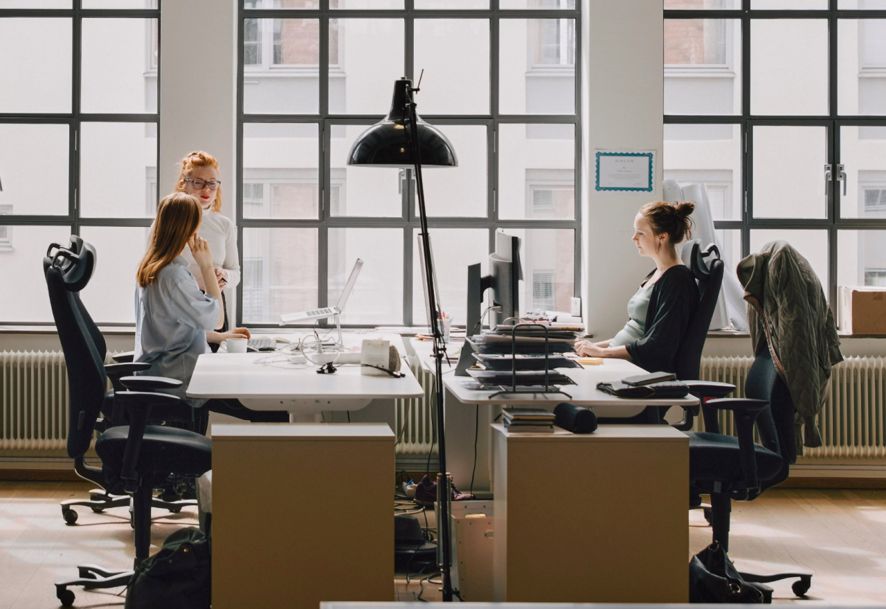 Two women discuss at one workstation while another woman works across from them in an office.