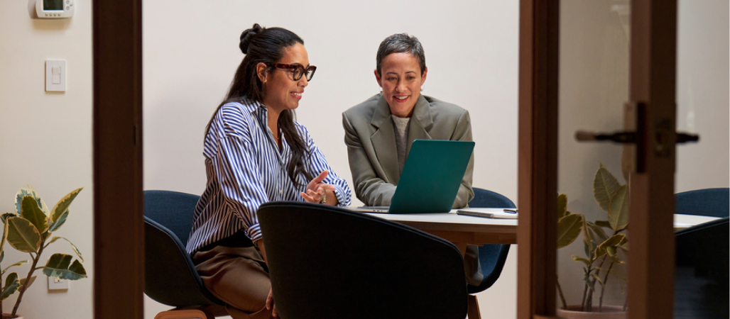 Image of two people sitting in an office space while looking at a laptop together.